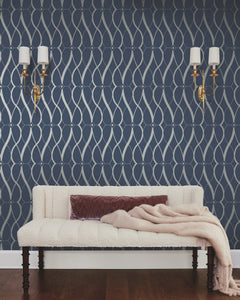 A white textured sofa with a mauve pillow and beige throw sits against a navy wall adorned with the Graceful Geo Wallpaper by York Wallcoverings. Two brass wall sconces with white shades are mounted above the sofa, and the room features a dark wood floor.