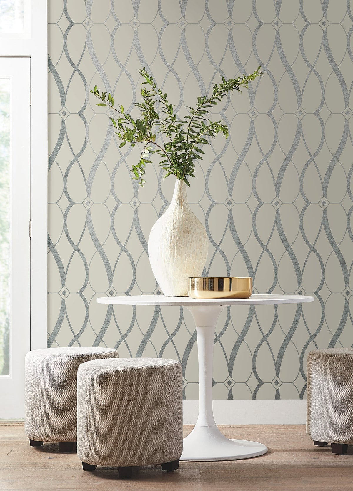 A white vase with green branches sits on a round white table next to a gold tray. Two round beige ottomans are nearby, with York Wallcoverings’ Graceful Geo Wallpaper and a window in the background.