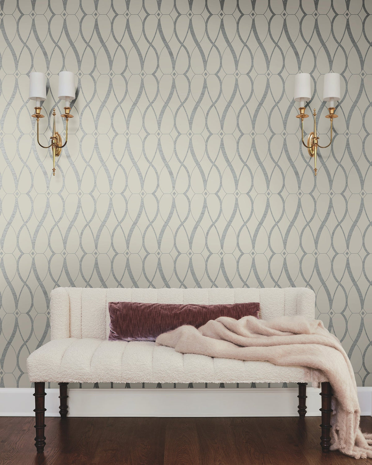 A cream-colored couch with a mauve pillow and beige throw blanket sits against a wall adorned with Graceful Geo Wallpaper by York Wallcoverings. Two gold wall sconces with white shades are mounted above the couch, and dark wood flooring is visible.