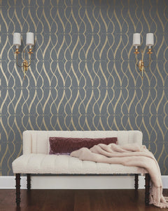 A cream-colored sofa with a mauve throw blanket sits against a wall adorned with Graceful Geo Wallpaper by York Wallcoverings in a dark gray and gold geometric pattern. Two brass wall sconces with white shades are mounted above the sofa.