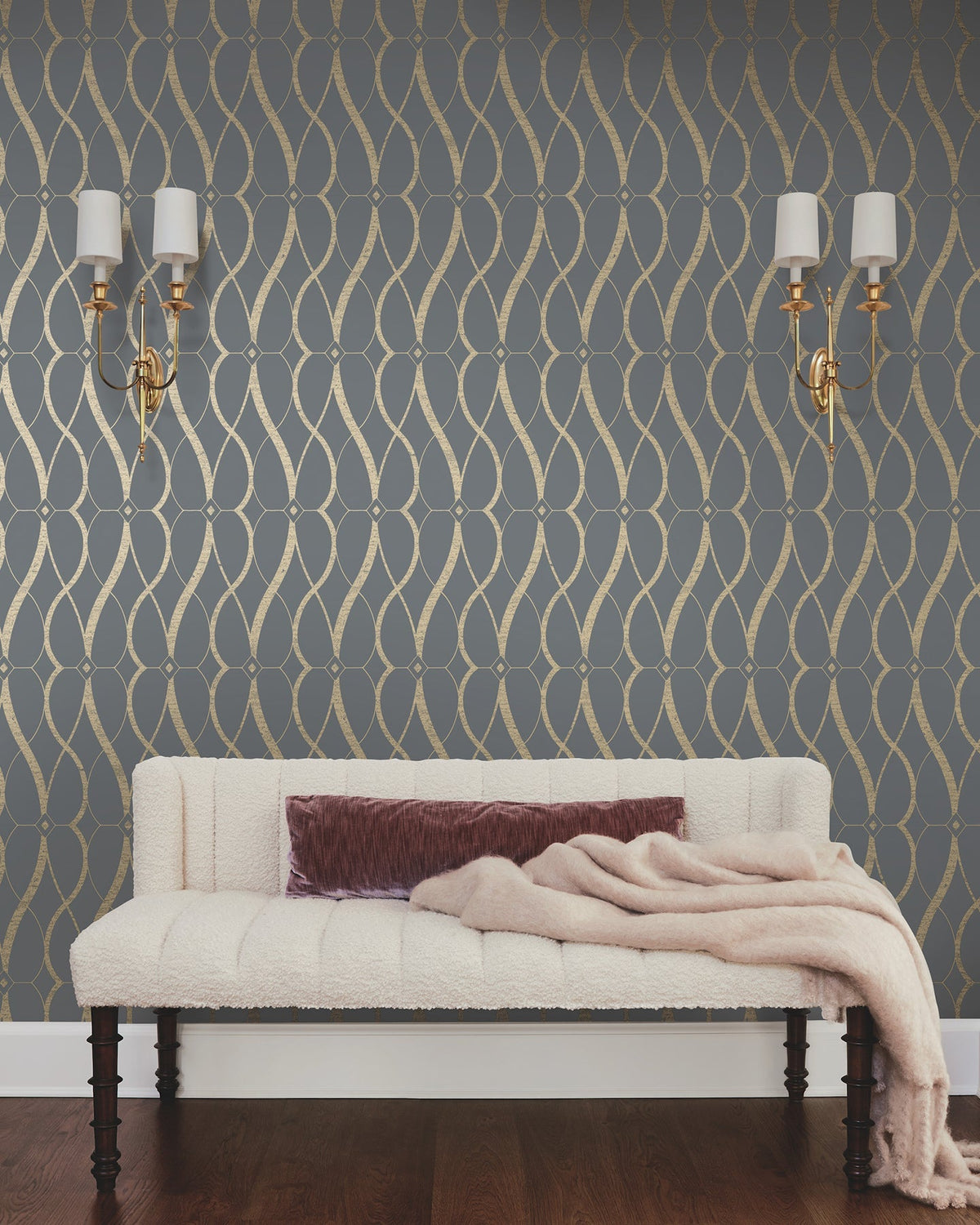 A cream-colored sofa with a mauve pillow and throw sits against a dark gray wall adorned with the Graceful Geo Wallpaper by York Wallcoverings. Two gold sconces with white shades are mounted above the sofa, and the floor is dark wood.