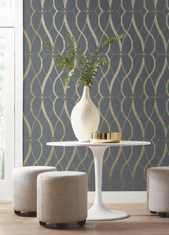 A white vase with green branches and a gold tray sit on a round white table, flanked by two beige stools, against a dark wall covered in Graceful Geo Wallpaper by York Wallcoverings. Light enters from a window on the left.