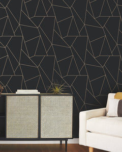 A modern living room featuring the Nazca Wallpaper by York Wallcoverings—a striking geometric black wall—paired with a light-colored couch, patterned sideboard, and decorative accents such as a spiky gold sculpture and stacked books.