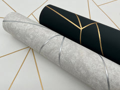 Two rolls of Nazca Wallpaper by York Wallcoverings rest on a surface, each featuring sleek, geometric lines. One roll showcases a gray background with shimmering silver accents, while the other displays a black base with striking gold lines. Both highlight modern, minimalist design aesthetics.