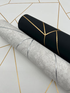 Two rolls of York Wallcoverings’ Nazca Wallpaper are displayed on a geometric-patterned surface: one roll is black with gold lines, the other is light gray with silver lines. The background also features intersecting gold lines.
