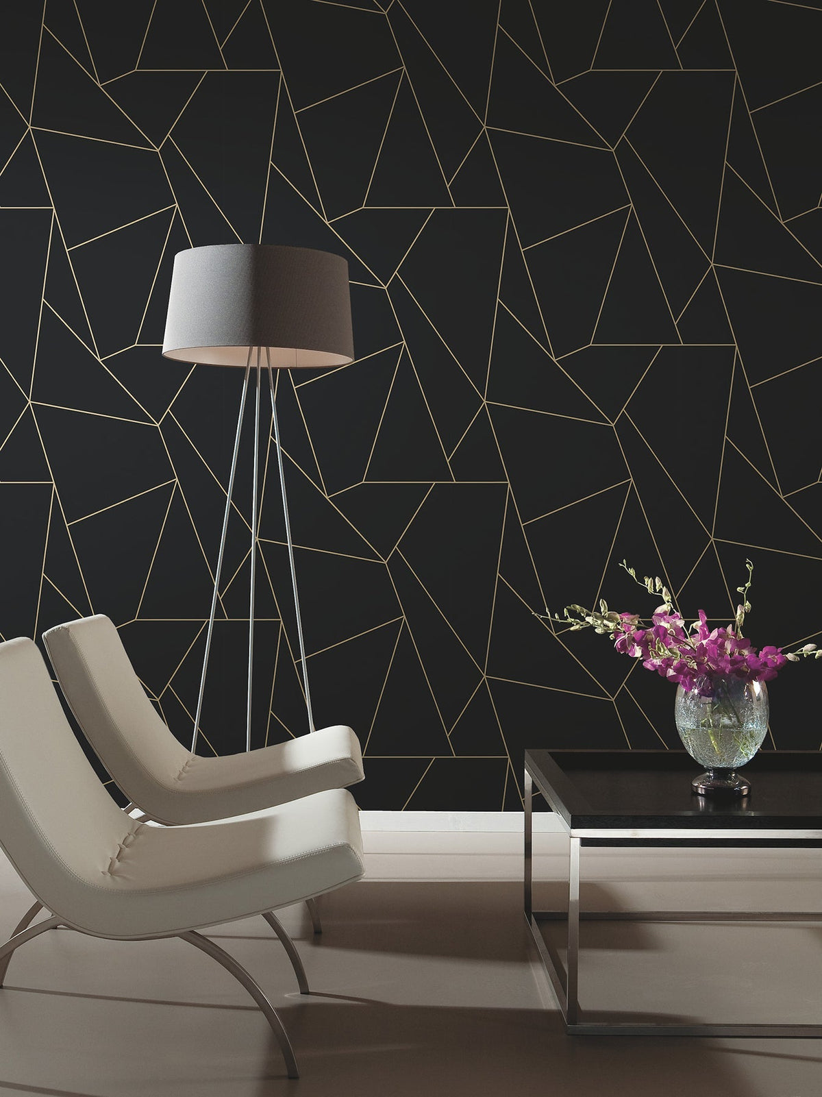 Two modern white chairs and a black lamp sit by a black coffee table with a glass vase of purple flowers, all set against York Wallcoverings’ Nazca Wallpaper in black with a gold geometric line pattern.