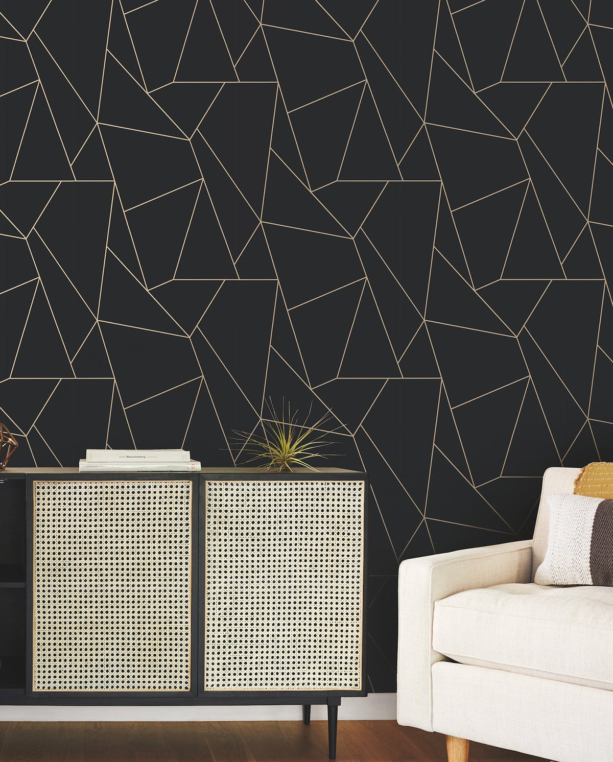 Modern living room with a beige sofa, a cabinet with rattan doors, a decorative plant, and a dark wall featuring the Nazca Wallpaper by York Wallcoverings with its geometric gold line pattern.