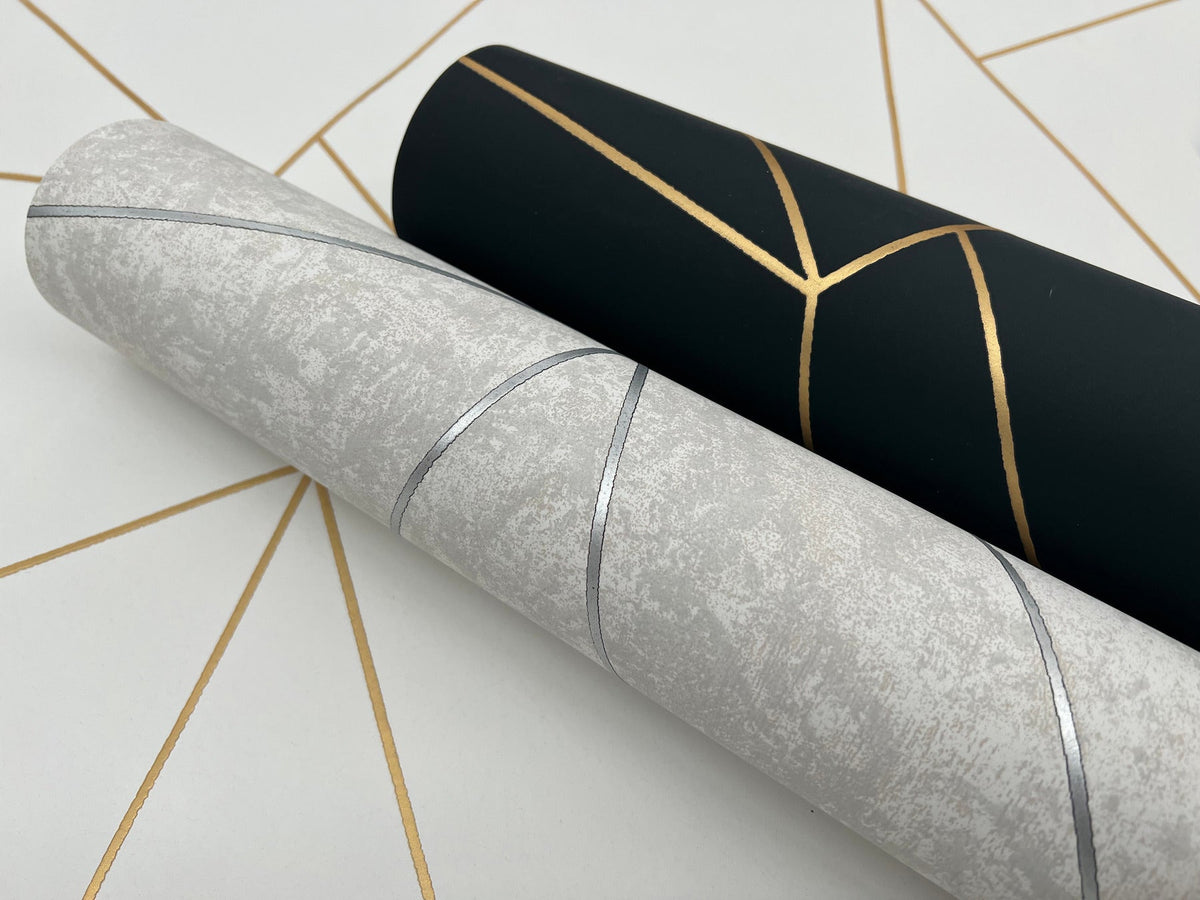 Two rolls of Nazca Wallpaper by York Wallcoverings are displayed on a white surface with gold geometric lines—one roll in black with gold accents, the other in light gray with subtle silver lines.