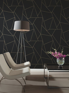 Two modern white chairs and a black floor lamp face a sleek black coffee table with a vase of purple flowers, set against a wall adorned with Nazca Wallpaper by York Wallcoverings, featuring a thin gold geometric pattern.