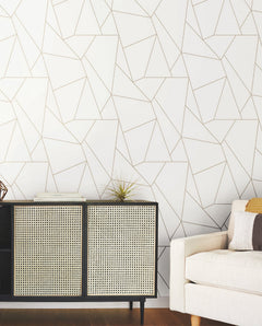 A modern living room with a beige sofa, a black and tan cabinet topped with a small plant and books, and a white wall adorned with the “Nazca Wallpaper” by York Wallcoverings featuring a gold geometric line pattern.