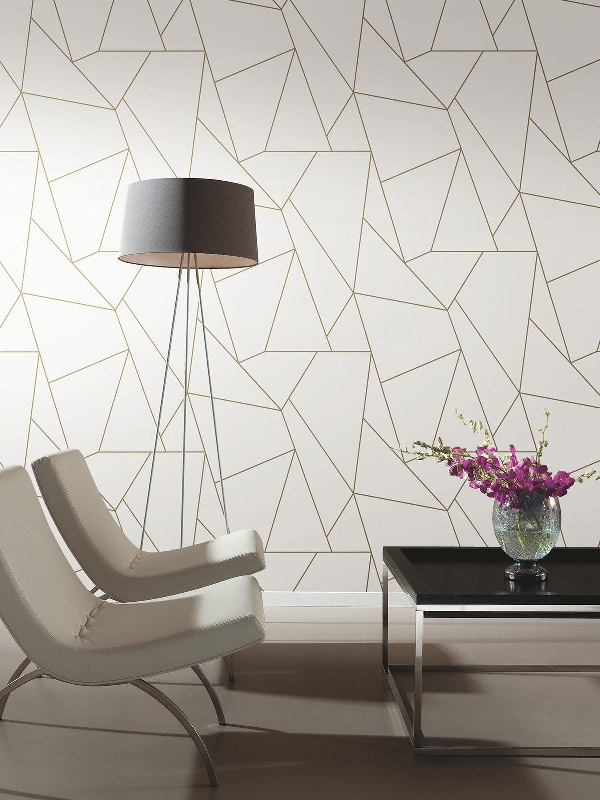 Two modern white chairs face a black coffee table with a glass vase of purple flowers. A tall floor lamp stands beside a wall covered in York Wallcoverings’ Nazca Wallpaper, featuring a geometric gold and white pattern.