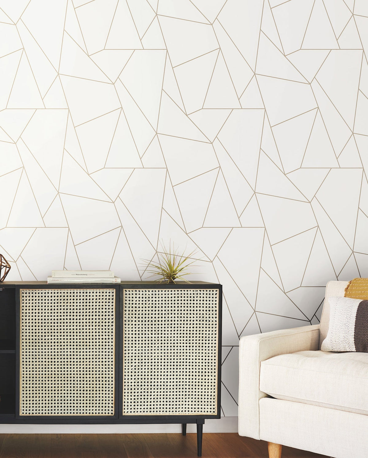 A modern room featuring a beige sofa and a black and wicker cabinet adorned with decor, set against a white wall covered in York Wallcoverings’ Nazca Wallpaper with a gold geometric line pattern. The flooring is wooden.