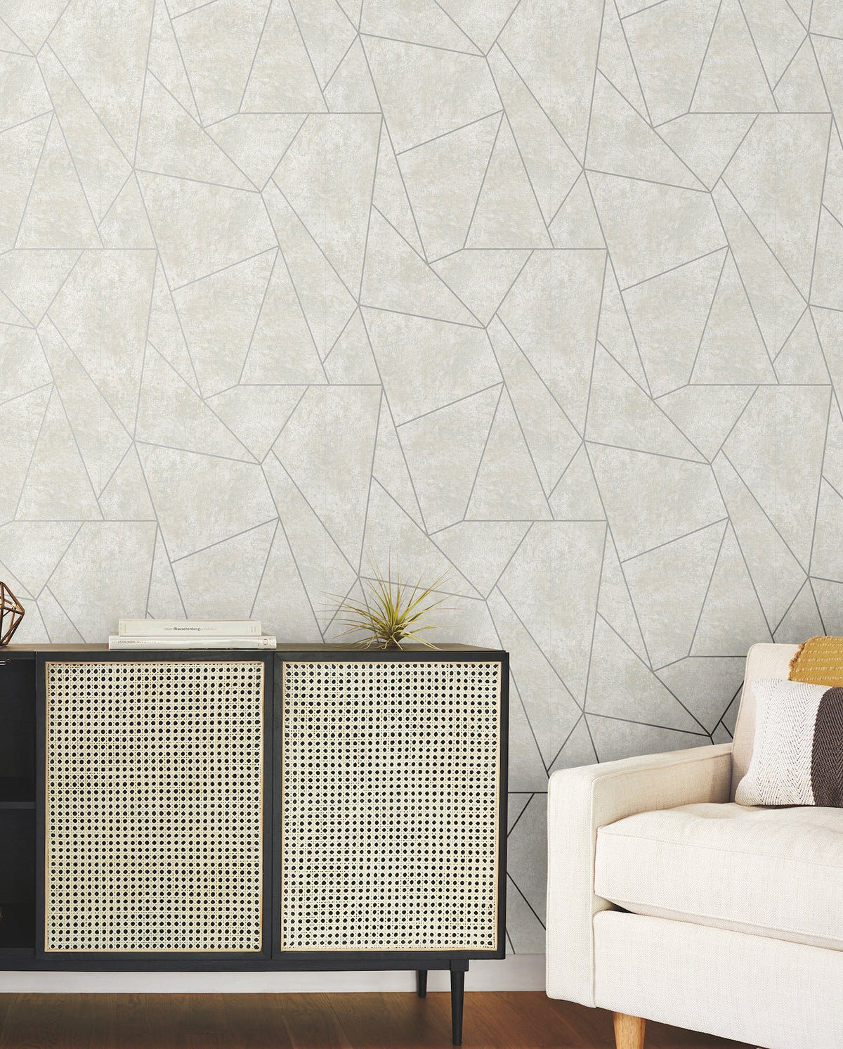 A modern living room featuring a beige sofa, a black and cane cabinet, books, a small plant, and York Wallcoverings’ Nazca Wallpaper in a light gray geometric pattern.