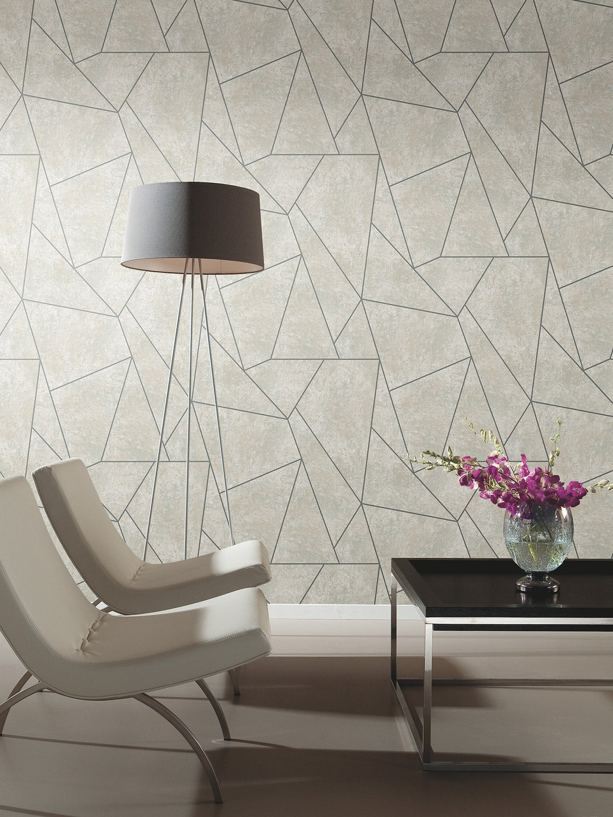 Two modern white chairs sit near a tall floor lamp and a black coffee table topped with a glass vase of purple flowers. Behind them, the wall is adorned with York Wallcoverings’ Nazca Wallpaper, featuring a beige geometric pattern with intersecting lines.