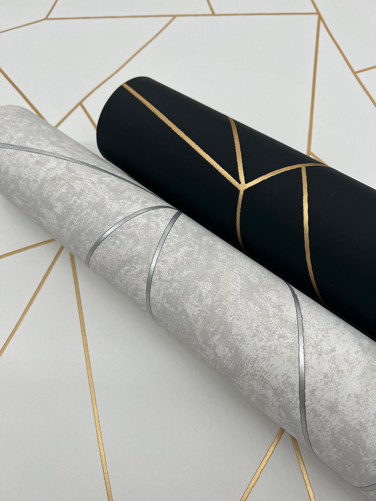 Two rolls of Nazca Wallpaper by York Wallcoverings—one in black with gold geometric lines and the other in light gray with silver lines—are displayed on a white surface decorated with a gold geometric pattern.