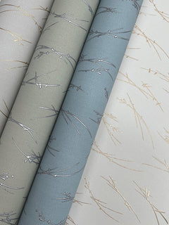 Three rolls of York Wallcoverings’ Sprigs Wallpaper in light gray, green-gray, and blue-gray shades, each featuring a textured metallic branch pattern in gold and silver tones, displayed side by side on a light background.