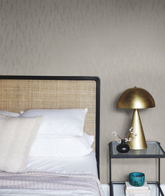 A modern bedroom featuring a rattan headboard, white pillows, a gray blanket, a brass table lamp, a small blue bowl, books, and a glass nightstand adorned with decorative branches. The background showcases York Wallcoverings’ Sprigs Wallpaper in a pale pattern.