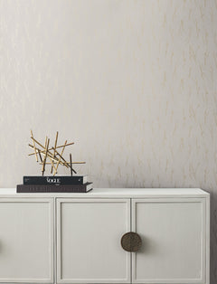 White cabinet with round handles, topped with stacked books, a magazine, and a modern gold abstract sculpture. The background features Sprigs Wallpaper by York Wallcoverings—a light design with a subtle gold botanical pattern.