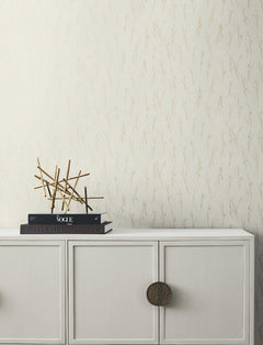 A white cabinet with round brass handles holds stacked books and a modern gold sculpture, set against York Wallcoverings’ Sprigs Wallpaper in a cream hue with a subtle branch pattern.