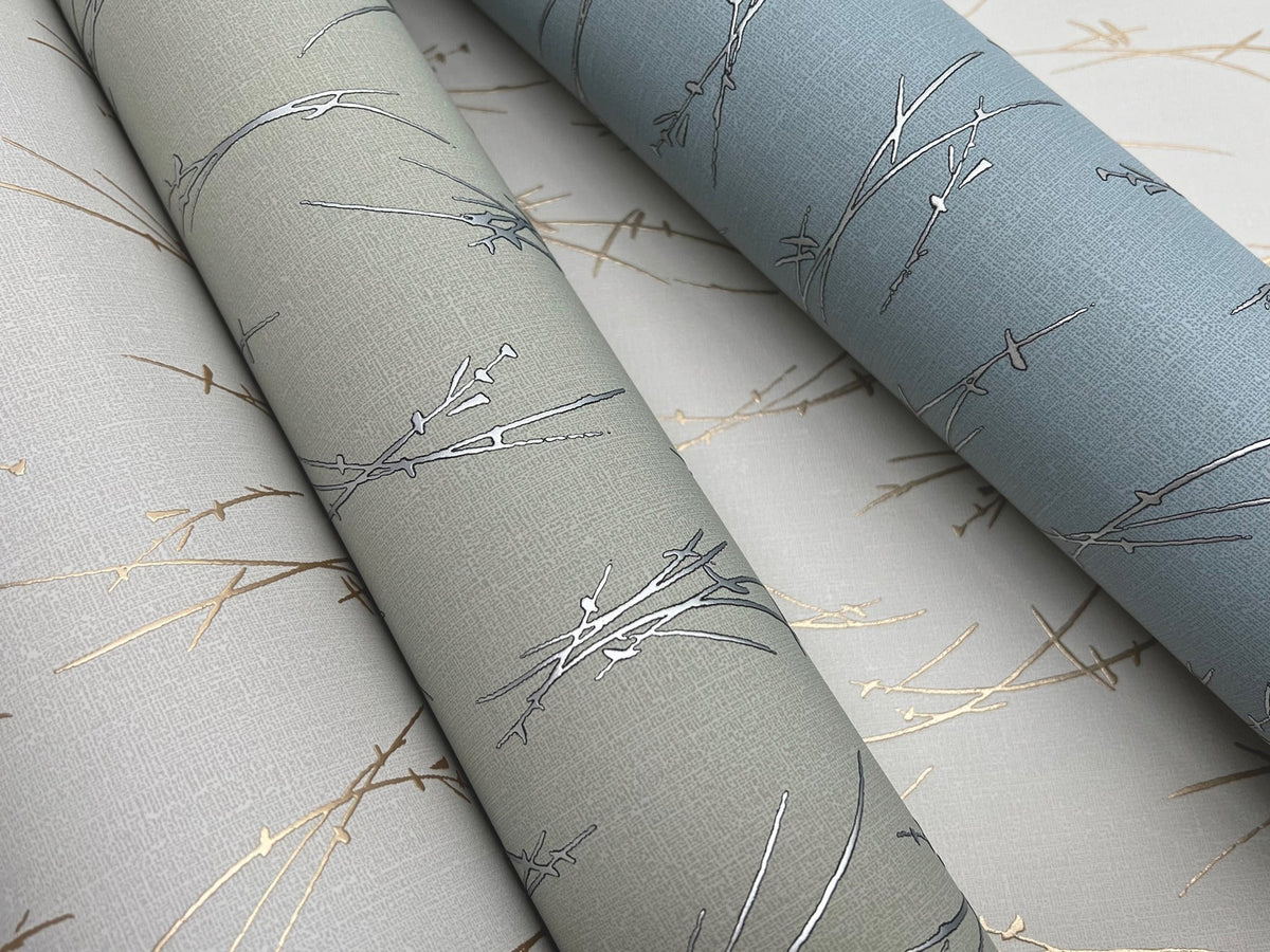 Sprigs Wallpaper by York Wallcoverings features three rolls in neutral tones—light beige, sage green, and dusty blue—each adorned with minimalist metallic branch patterns in silver or gold. The design is subtly embossed for added texture.