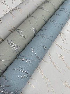 Three rolls of York Wallcoverings’ Sprigs Wallpaper in muted beige, green, and blue shades, each featuring a delicate metallic branch pattern, are placed side by side on a matching patterned surface.