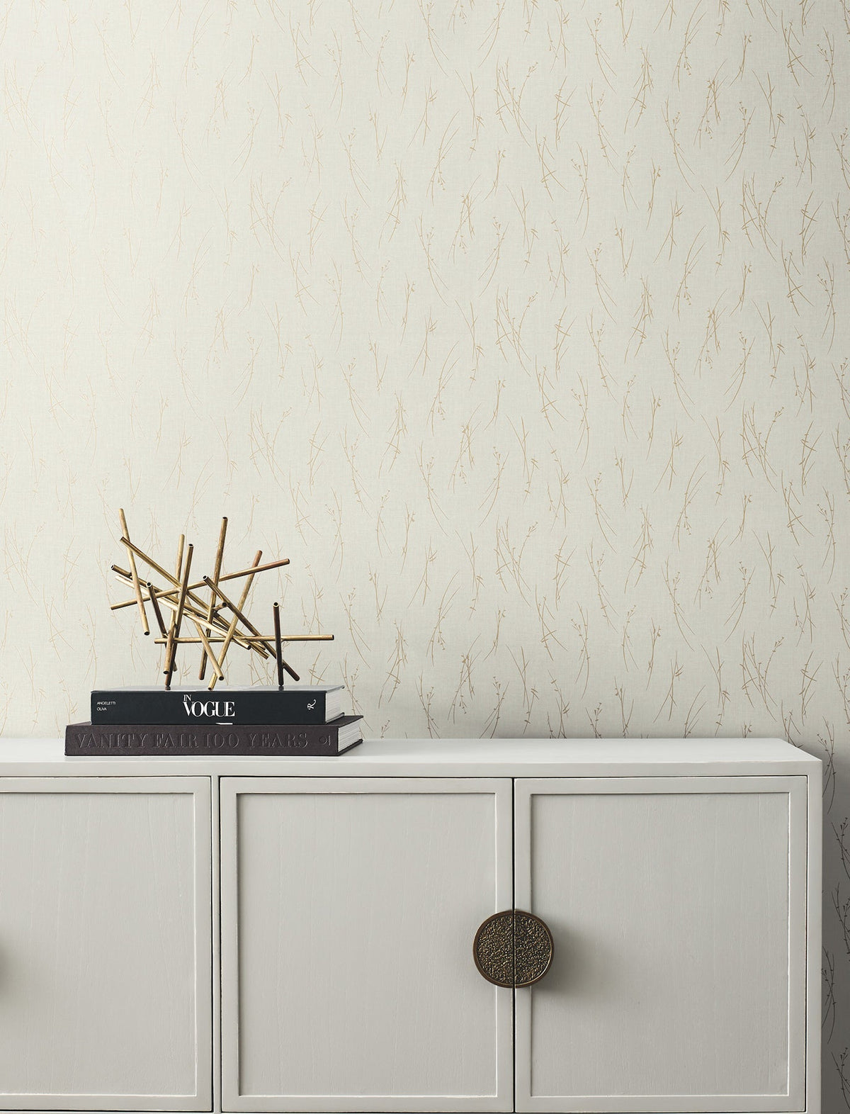Minimalist decor scene featuring a light-colored cabinet with round bronze handle, stacked books, and a gold abstract sculpture, set against York Wallcoverings’ Sprigs Wallpaper in beige with a subtle branch motif.