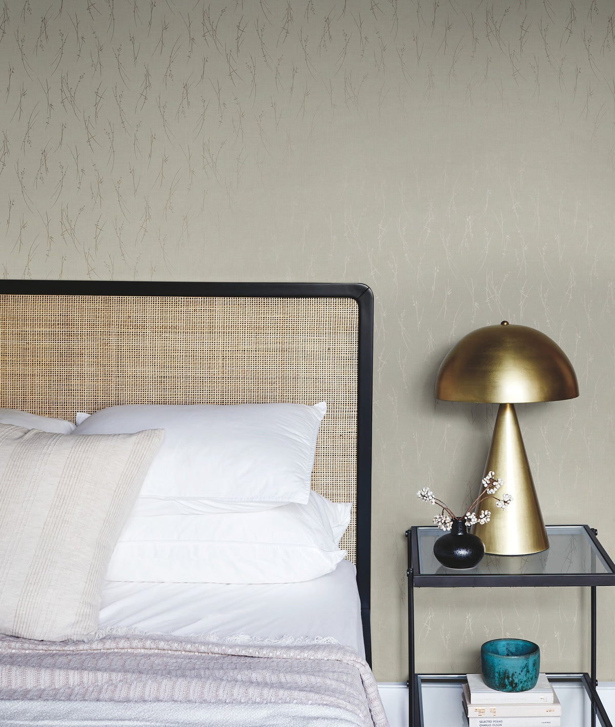 A modern bedroom featuring York Wallcoverings’ Sprigs Wallpaper as the backdrop, with a rattan headboard, white pillows, a gold table lamp, a small vase of flowers, a blue bowl, and books arranged on a glass nightstand.