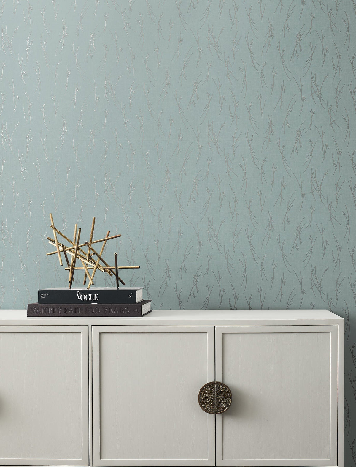 A white cabinet with round handles sits against a blue wall adorned with York Wallcoverings’ Sprigs Wallpaper. On top are books and a modern gold decorative sculpture.