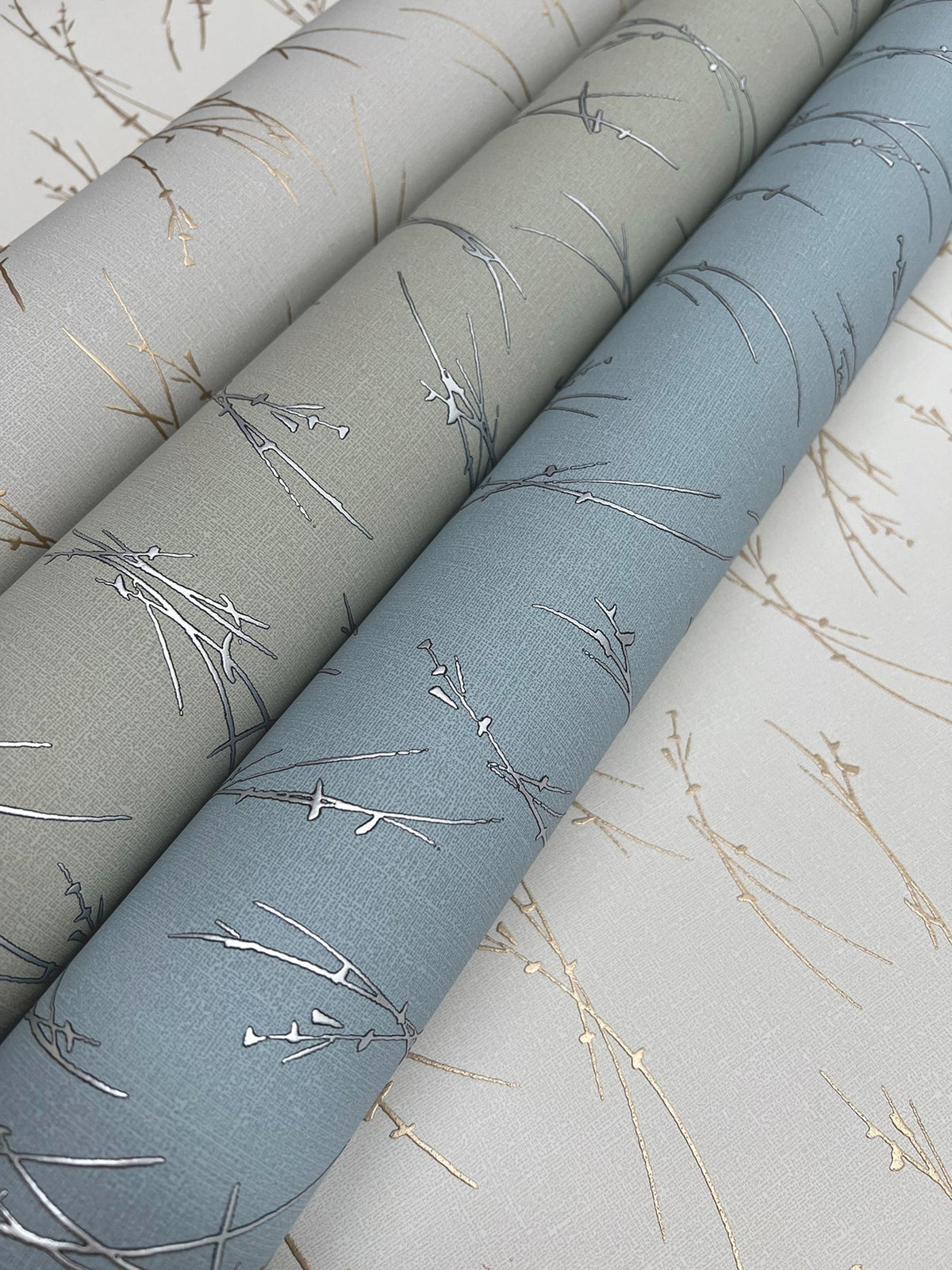 Three rolls of York Wallcoverings’ Sprigs Wallpaper in light gray, sage green, and blue—each featuring a delicate metallic branch pattern—are displayed side by side on a matching background.