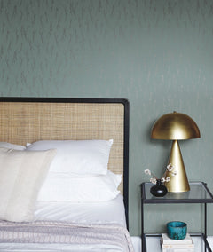 A bed with white and beige pillows sits next to a black nightstand topped with a gold lamp, blue cup, small branches in a vase, and books, set against York Wallcoverings’ Sprigs Wallpaper in green.