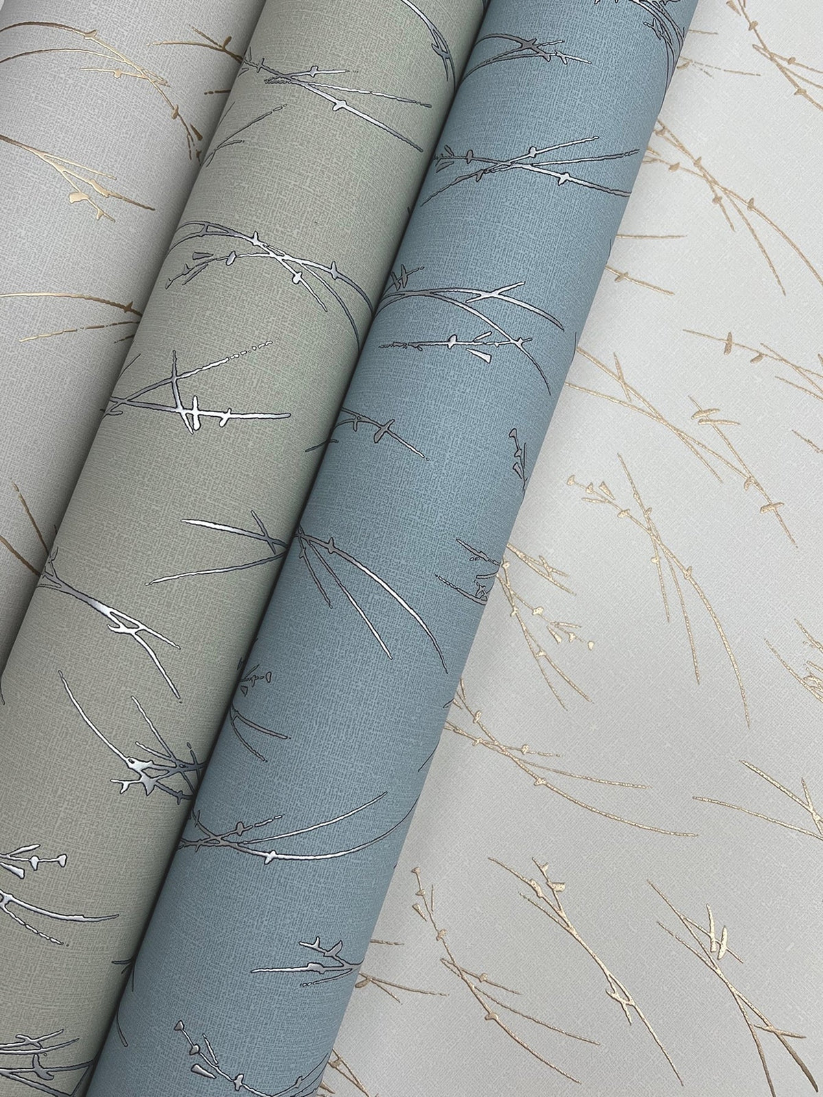 Three rolls of York Wallcoverings’ Sprigs Wallpaper in gray, blue, and white—each featuring delicate metallic branch patterns—are arranged diagonally on a matching patterned background.