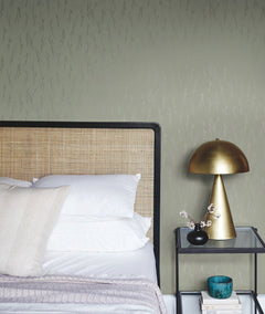 A neatly made bed with white pillows and a beige headboard stands beside a black metal nightstand holding a gold lamp, a small vase with branches, a teal bowl, and stacked books, all set against a green textured wall covered in the Sprigs Wallpaper by York Wallcoverings.