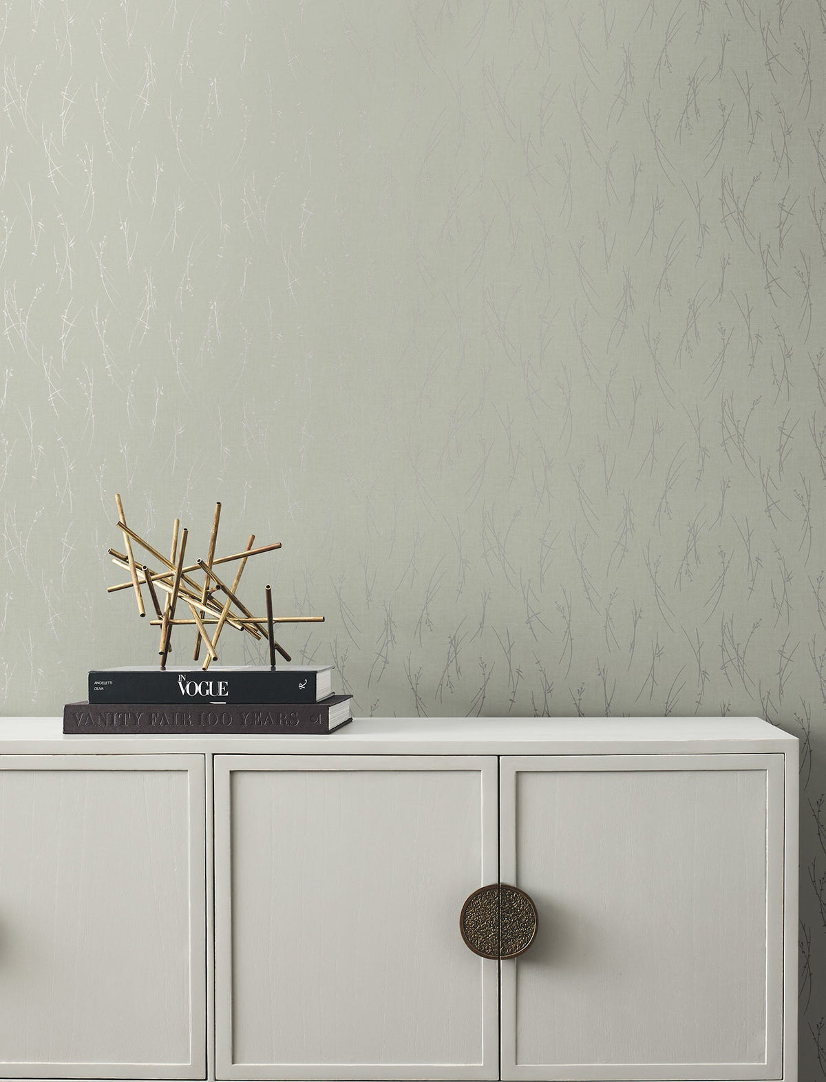 A minimalist white cabinet with round handles sits against a wall adorned in York Wallcoverings’ Sprigs Wallpaper. On top, stacked books and a modern gold decorative sculpture complete the scene.