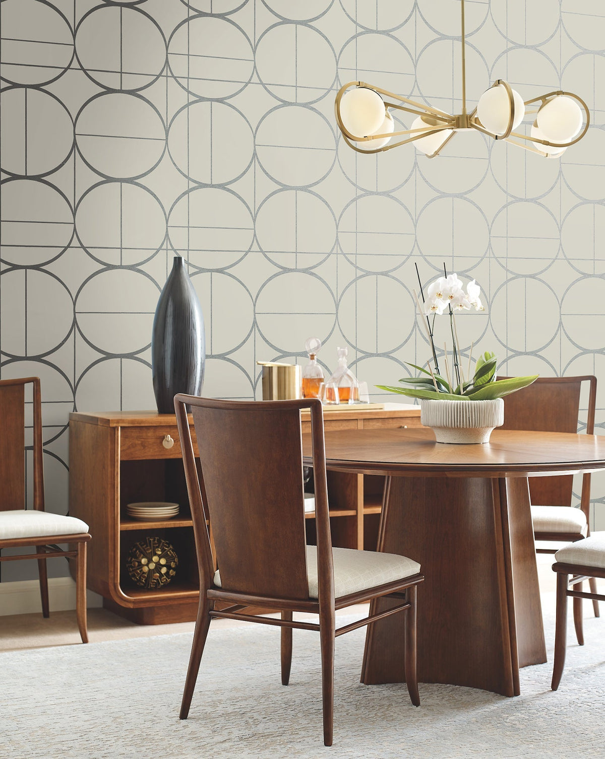 Mid-century modern dining room with a wooden table, four chairs, a sideboard, gold chandelier, and decorative items including a vase, bowl with white orchid, and gold canister. The walls feature Sun Circles Wallpaper by York Wallcoverings.