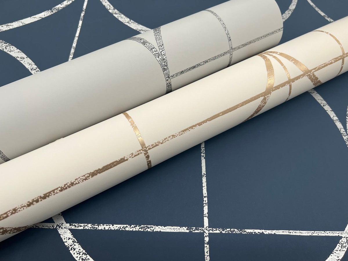Two rolls of wallpaper—one white with metallic silver lines and one cream with metallic gold lines—are displayed on top of the Sun Circles Wallpaper by York Wallcoverings, which features a navy background with a white geometric pattern.