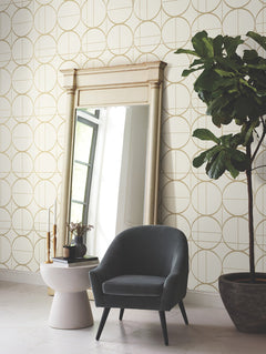 A gray armchair sits by a small white table with books and candles, next to a tall mirror and a large potted plant, against a wall adorned with York Wallcoverings’ Sun Circles Wallpaper.