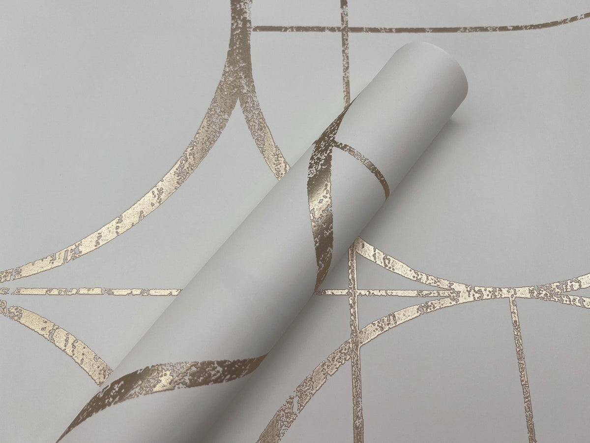 A roll of York Wallcoverings Sun Circles Wallpaper, featuring a white background with a metallic gold geometric pattern of intersecting curved and straight lines, lies partially unrolled atop matching wallpaper.