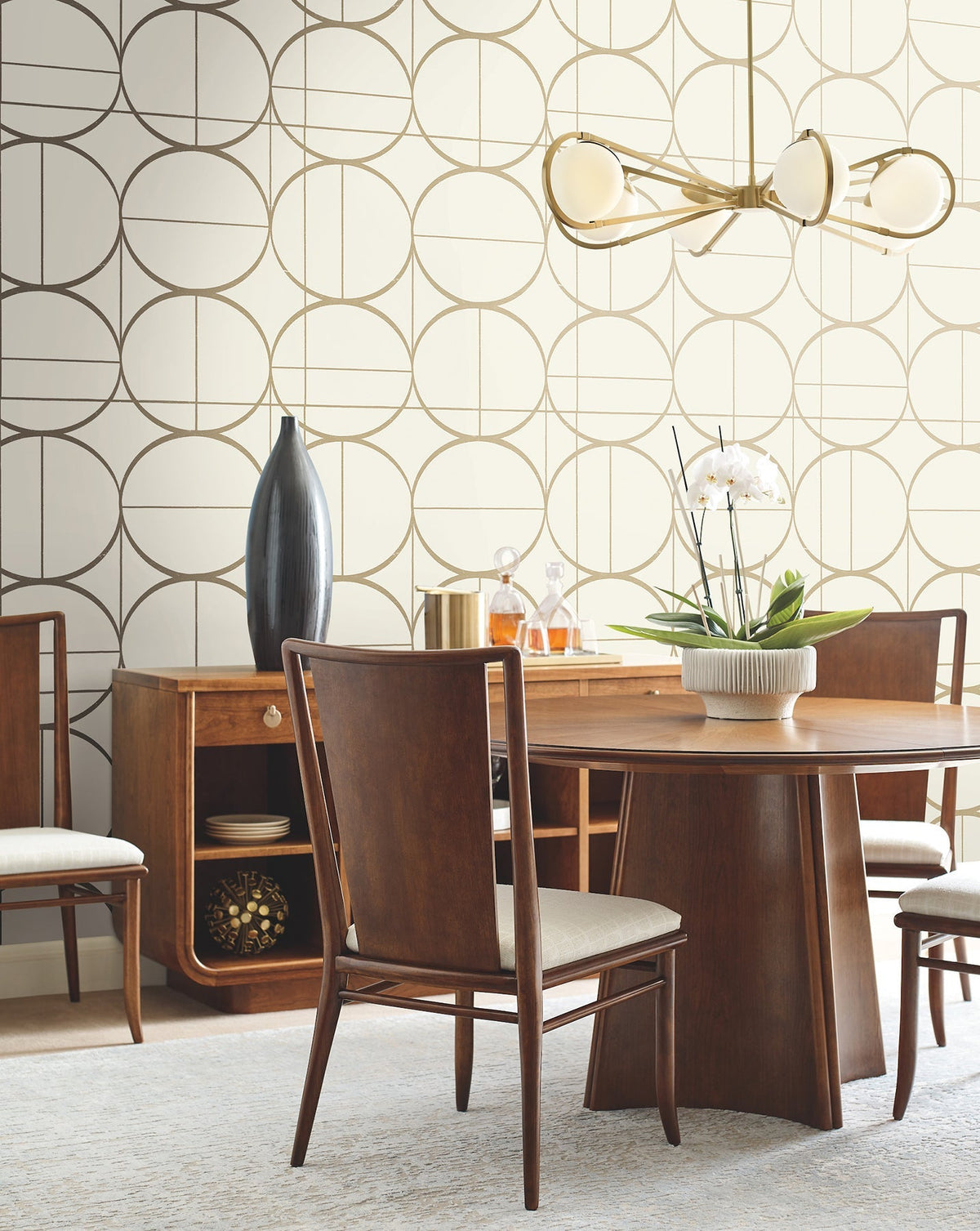 Mid-century modern dining room with wooden furniture, a round table, and York Wallcoverings’ Sun Circles Wallpaper. A gold chandelier hangs overhead, while a white orchid centerpiece decorates the table. A tall black vase and gold accents sit on a sideboard.