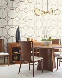 Mid-century modern dining room with wooden furniture, a round table, and York Wallcoverings’ Sun Circles Wallpaper. A gold chandelier hangs overhead, while a white orchid centerpiece decorates the table. A tall black vase and gold accents sit on a sideboard.
