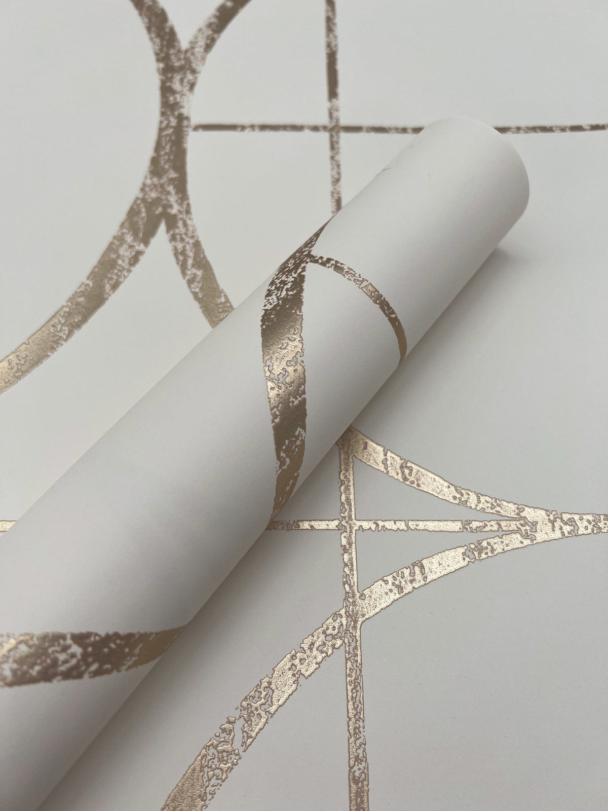 A partially unrolled roll of Sun Circles Wallpaper by York Wallcoverings in white, featuring shiny gold geometric patterns, displayed on a matching background.