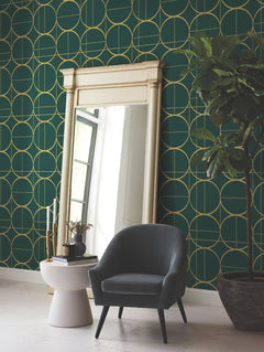A modern room featuring York Wallcoverings’ Sun Circles Wallpaper in green with a gold geometric pattern, complemented by a large ornate mirror, a small round side table with decor, a dark gray armchair, and a potted plant.