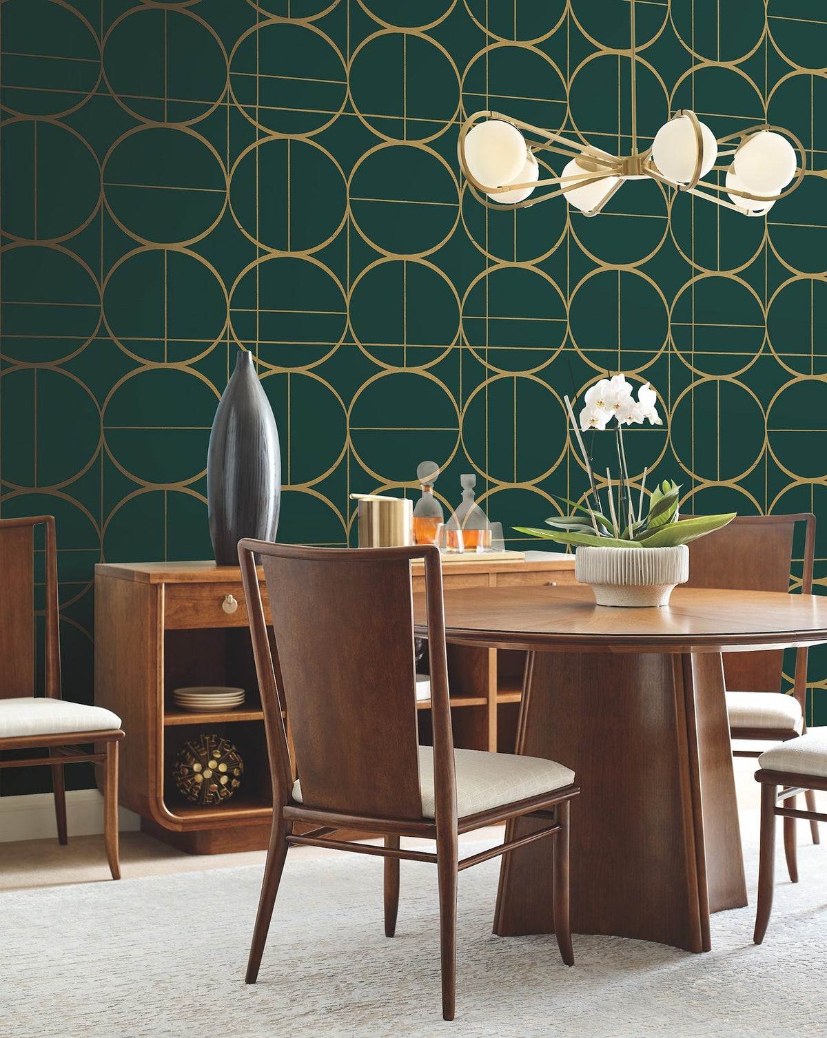 A modern dining room featuring York Wallcoverings’ Sun Circles Wallpaper in dark green and gold geometric patterns, paired with a wooden table and chairs, a sideboard adorned with decor items, and a white orchid centerpiece beneath a contemporary light fixture.