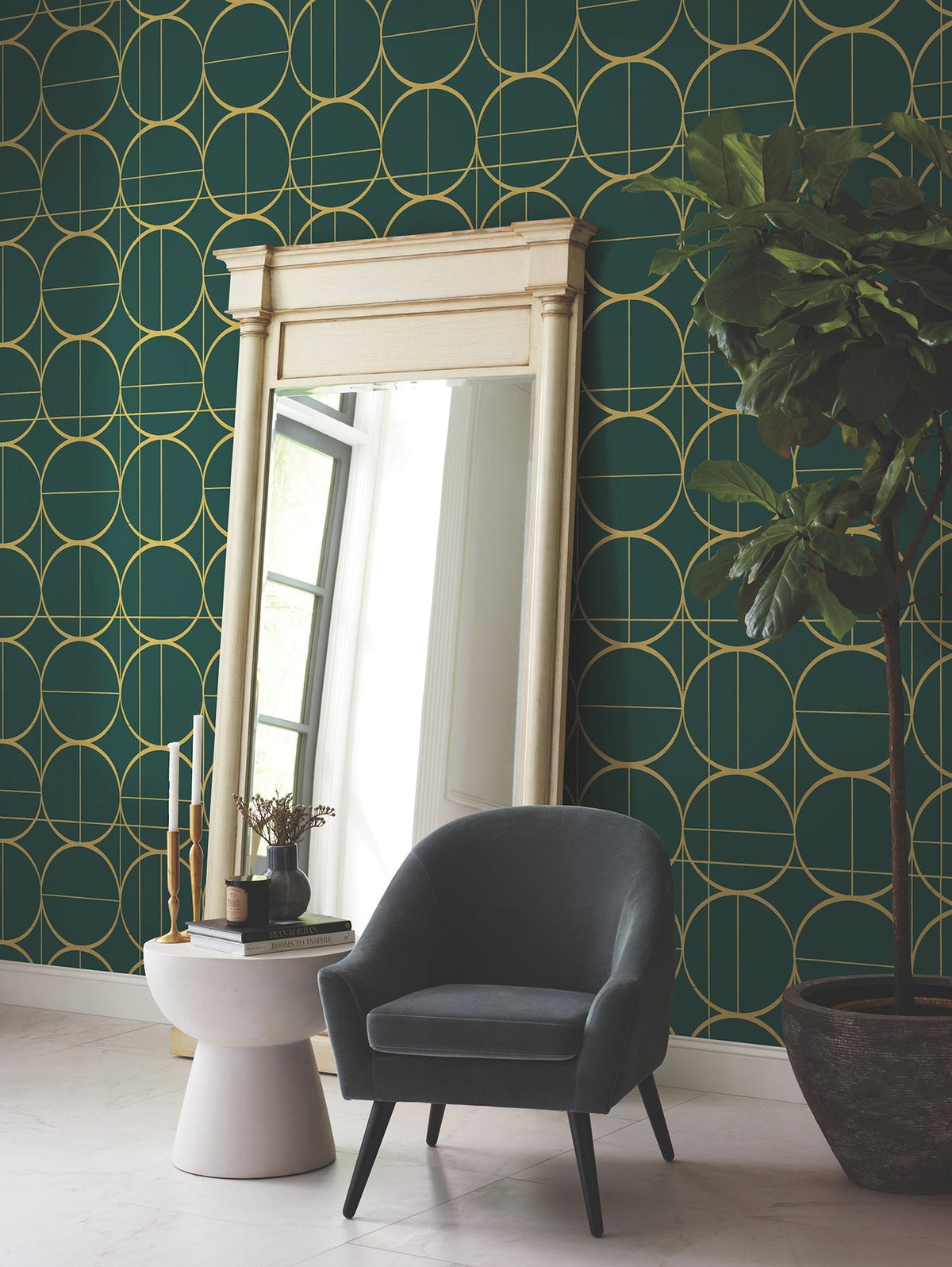 A large mirror with a cream frame stands against a green wall adorned with York Wallcoverings’ Sun Circles Wallpaper in gold geometric patterns. A gray armchair sits nearby, next to a round side table with decor, and a tall potted plant is on the right.