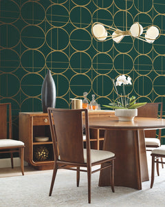A mid-century modern dining room featuring a wooden table and matching chairs, highlighted by York Wallcoverings’ Sun Circles Wallpaper in green and gold geometric patterns. The space is completed with a white orchid centerpiece, a sideboard adorned with vases, and a modern chandelier showcasing round white lights.