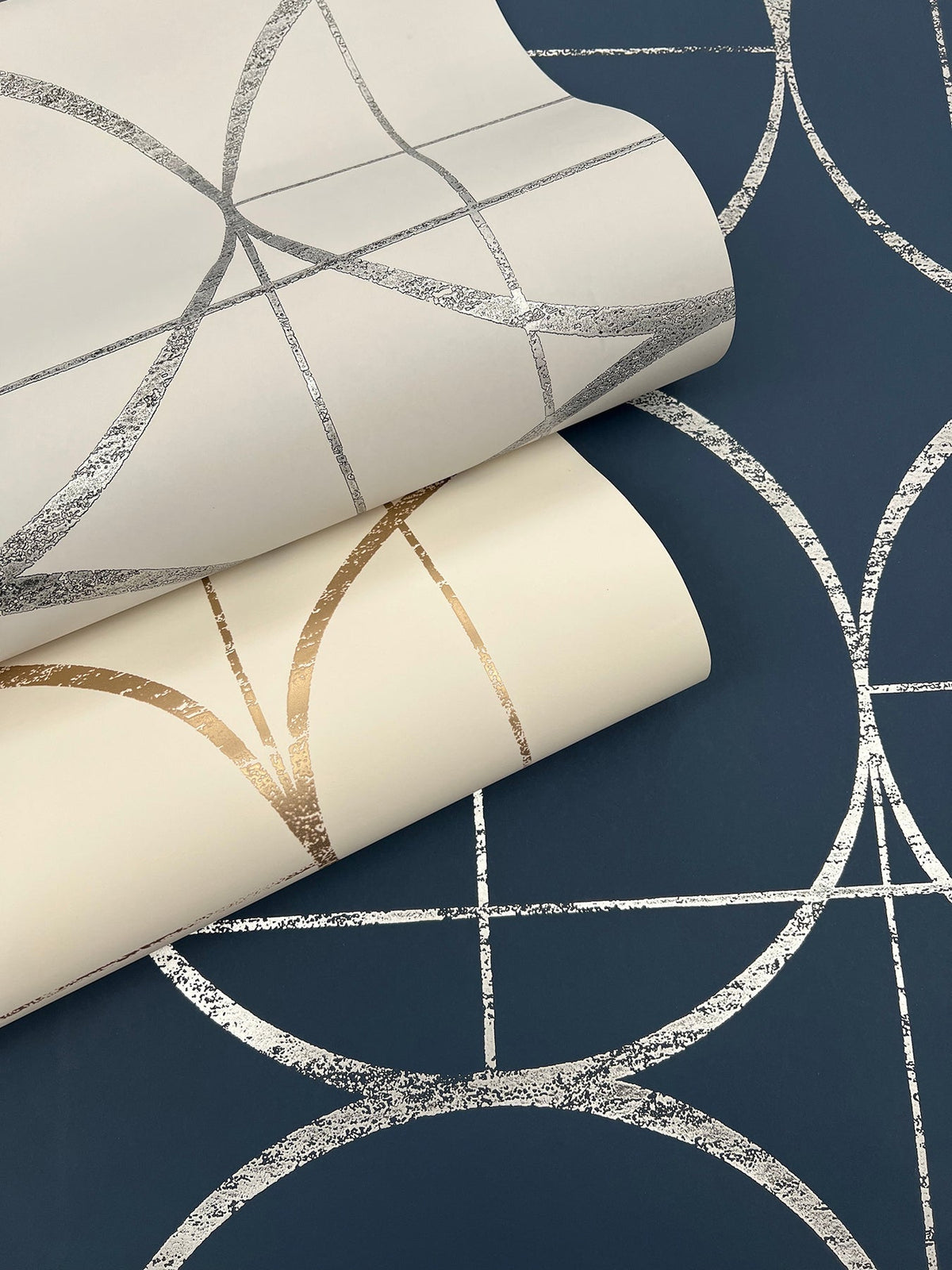 Two rolls of York Wallcoverings Sun Circles Wallpaper, one in white with gold geometric lines and the other in white with silver geometric lines, are displayed on a blue surface featuring a similar silver geometric pattern.