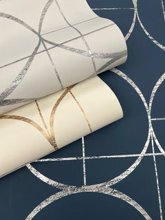 Two rolls of York Wallcoverings Sun Circles Wallpaper, one in white with gold geometric lines and the other in white with silver geometric lines, are displayed on a blue surface featuring a similar silver geometric pattern.
