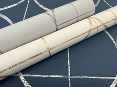 Two rolls of Sun Circles Wallpaper by York Wallcoverings—one featuring silver lines and the other gold—are displayed on a dark blue surface with a coordinating abstract lined pattern.