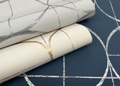 Two rolls of York Wallcoverings’ Sun Circles Wallpaper, featuring metallic geometric line patterns in silver and gold, are placed on top of a dark blue wallpaper displaying a similar silver geometric design.