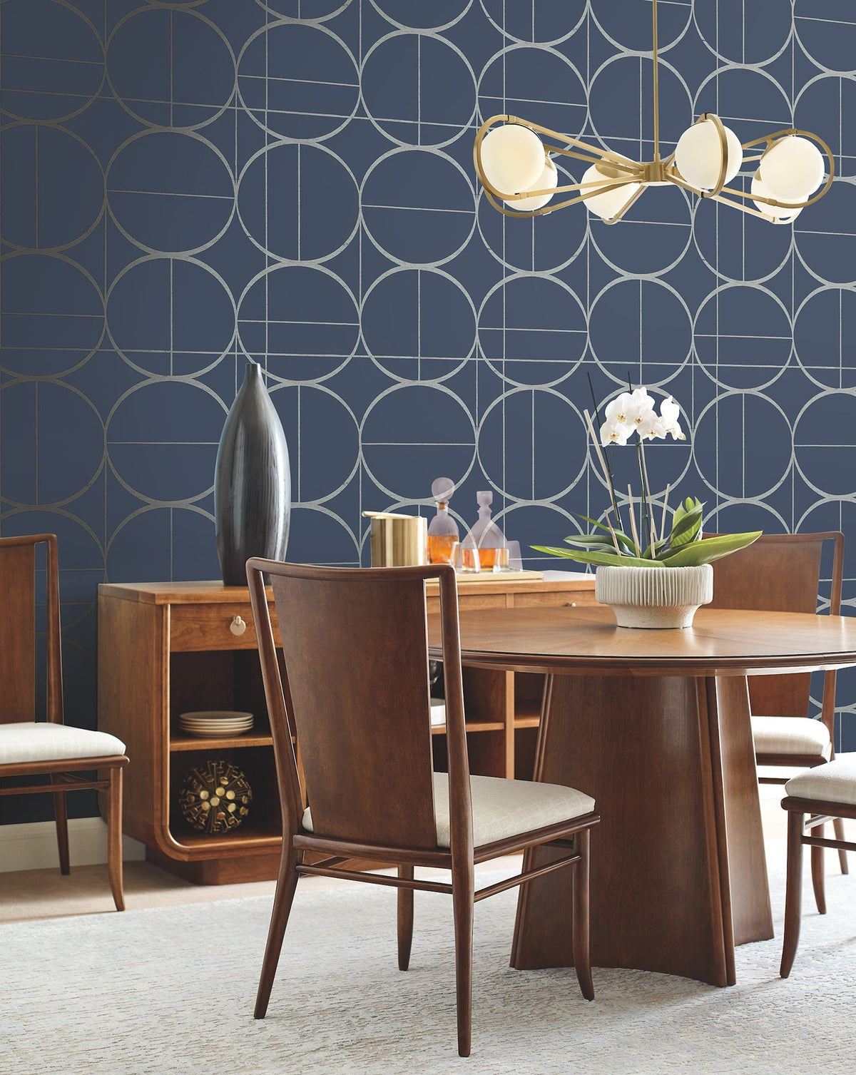 A modern dining room featuring a wooden table, four chairs, a sideboard, contemporary light fixture, a white orchid centerpiece, and York Wallcoverings’ Sun Circles Wallpaper in navy with geometric patterns.