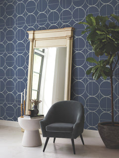 A tall mirror leans against a blue wall adorned with the Sun Circles Wallpaper by York Wallcoverings, featuring a geometric circle pattern. Nearby are a gray armchair, a white side table with decor items, and a large potted plant on a white floor.